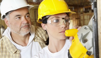 Female worker cutting wood with a power saw while male worker standing behind